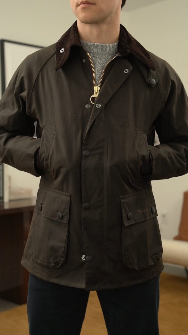 A man wearing a dark brown waxed jacket with a corduroy collar and gold zipper.