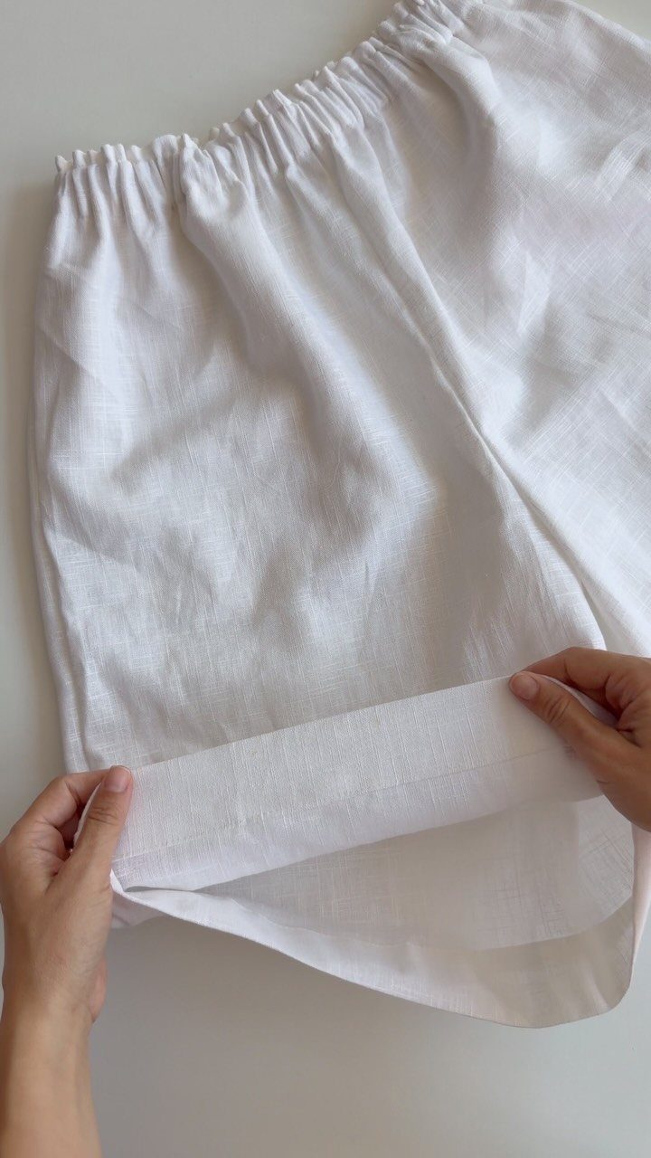 Hands holding up a pair of white linen shorts with an elastic waistband.
