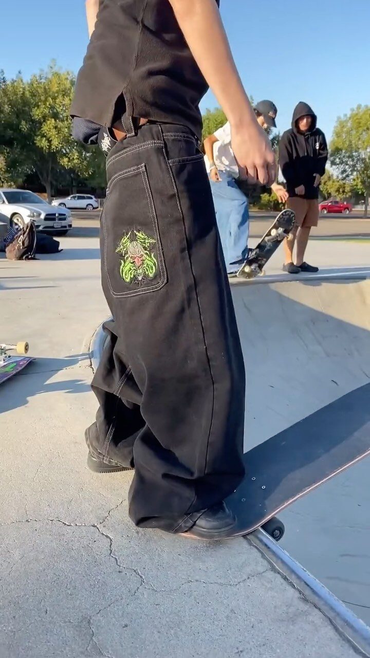 A person in black baggy pants stands on a skateboard at an outdoor concrete skatepark.