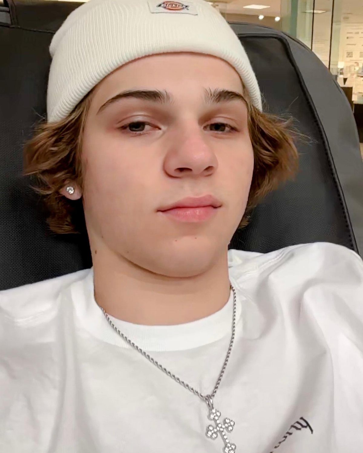 A young man wearing a white ribbed beanie and a silver cross necklace.