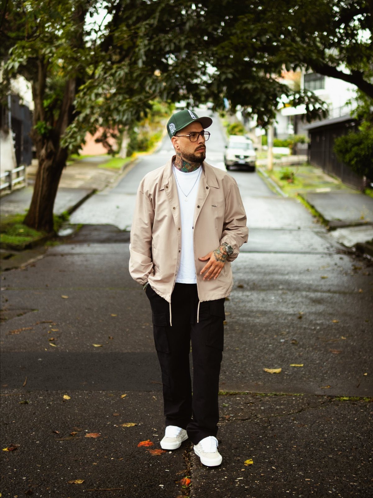 A man in skater fashion standing on a tree-lined residential street wearing a tan jacket and black pants.