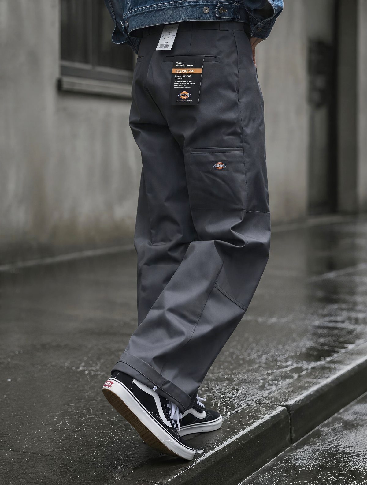 Person wearing baggy grey trousers and black sneakers standing on a wet urban sidewalk.
