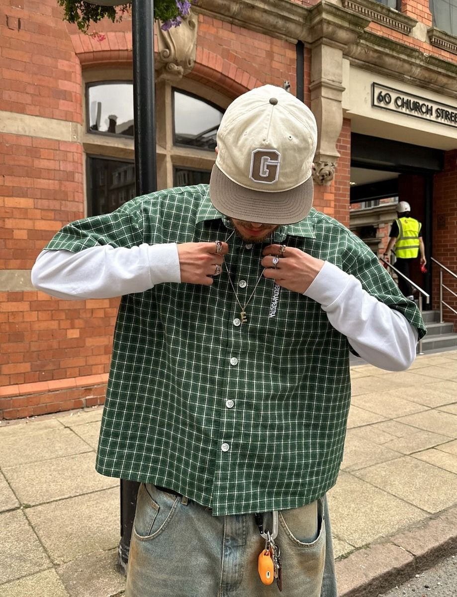 Man wearing a green checkered shirt layered over a white long-sleeve shirt and light blue jeans.
