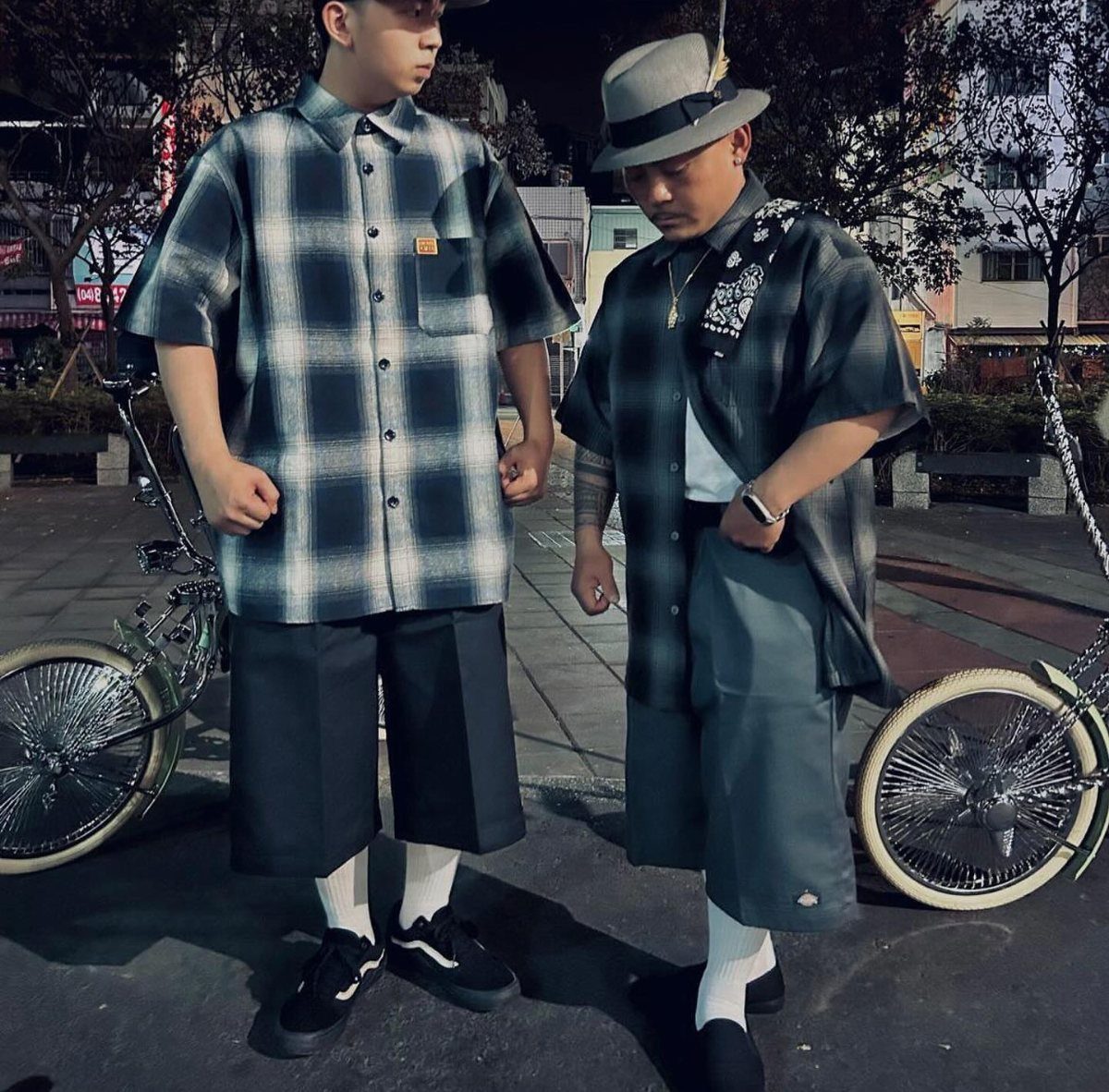 Two men posing in short-sleeved plaid shirts and dark shorts on a city street at night.