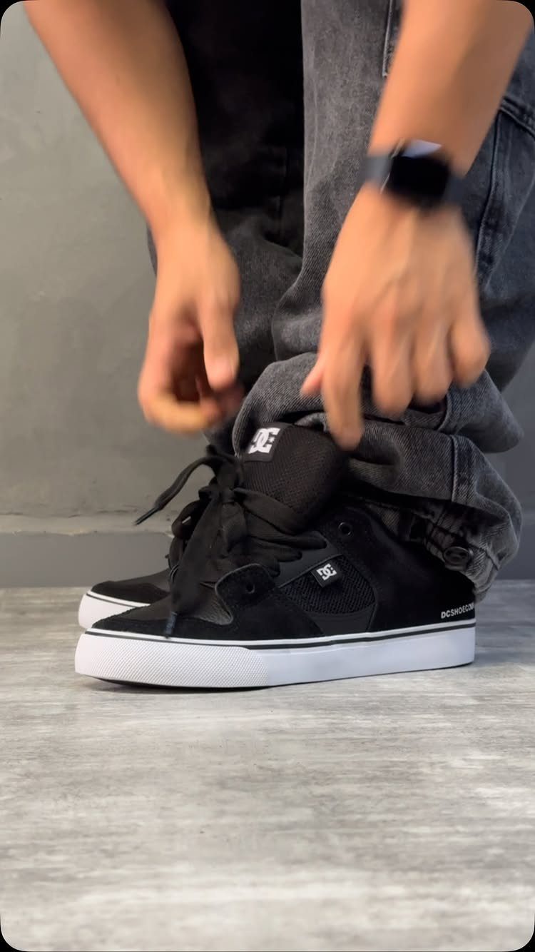 Hands tying the black laces of high-top DC skate shoes worn with grey jeans.