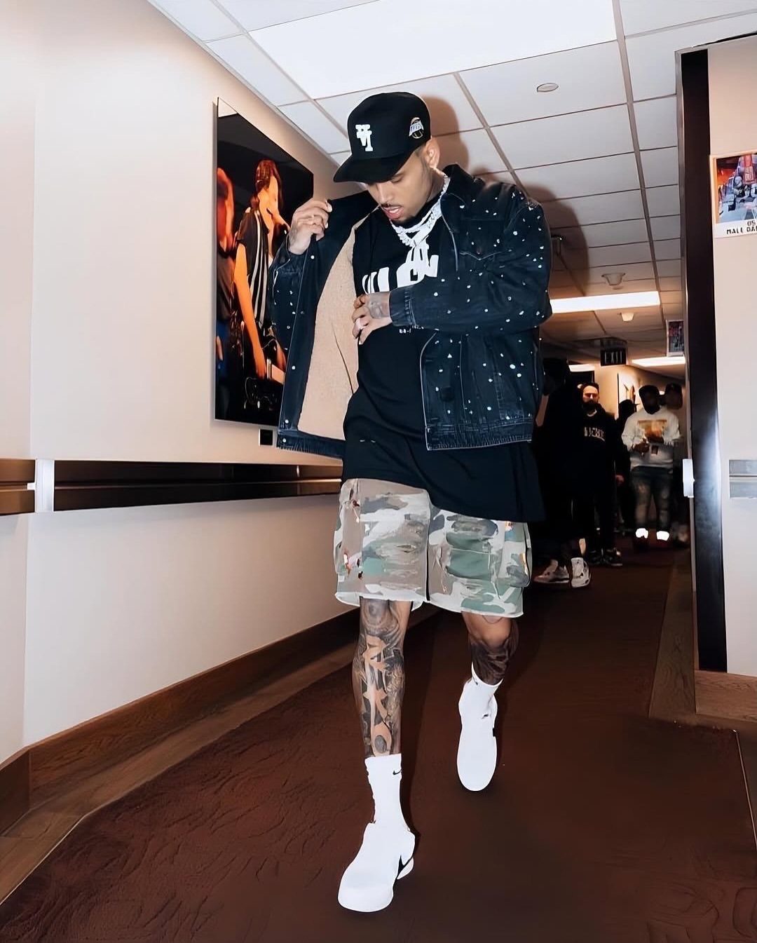 A man in a black denim jacket, camouflage shorts, and white sneakers walking down a hallway.