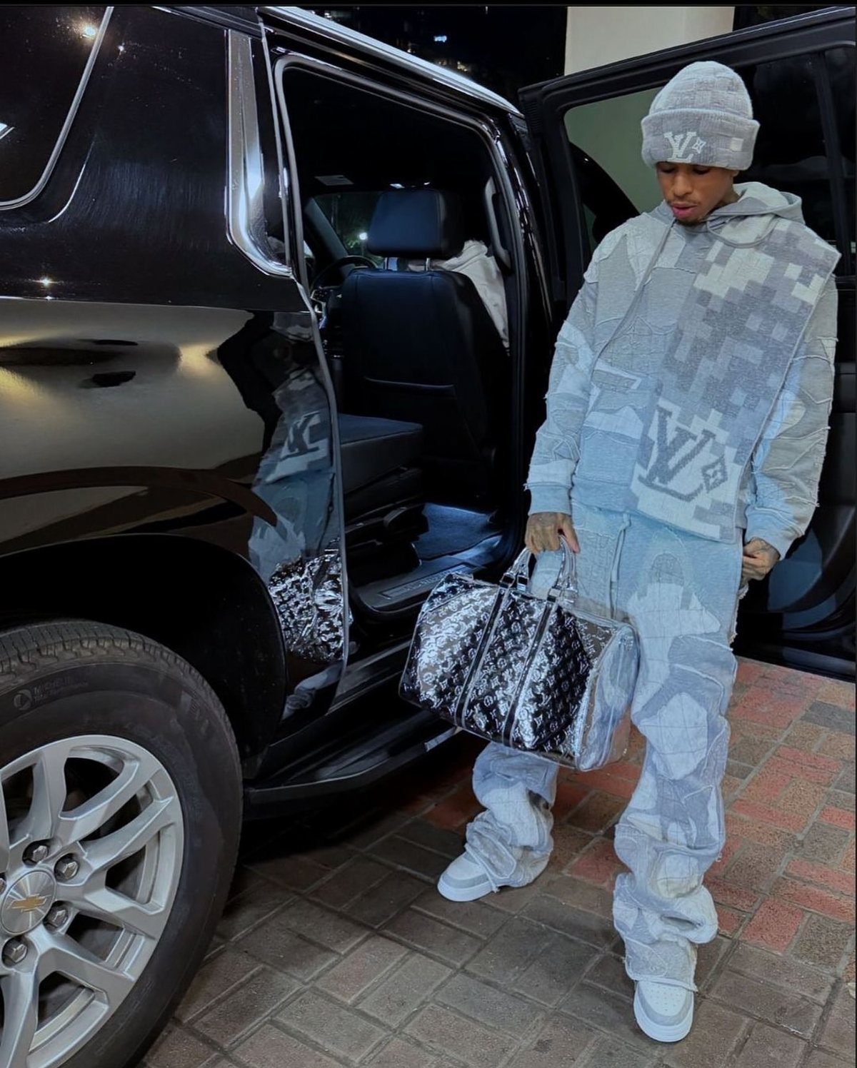 A man in a patterned light blue tracksuit stands by a black SUV holding a designer bag.