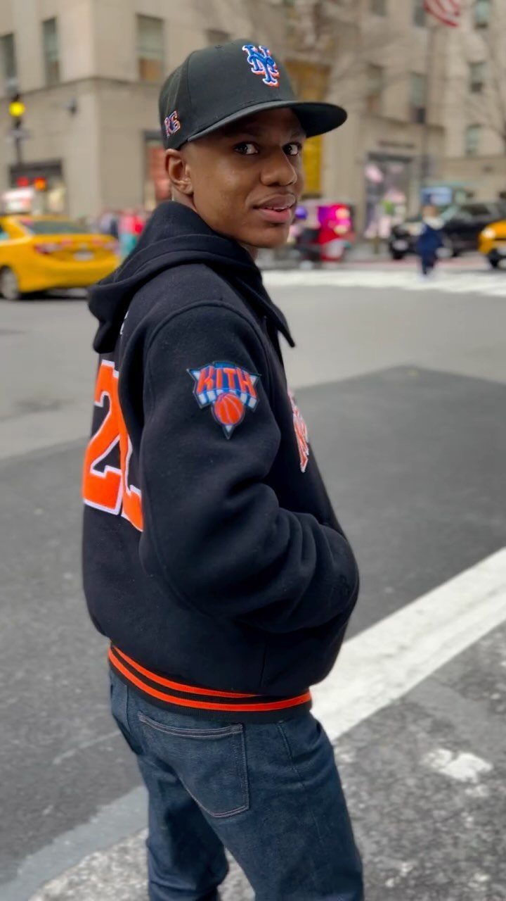 A young man wearing a black KITH hoodie and a baseball cap on a city street.