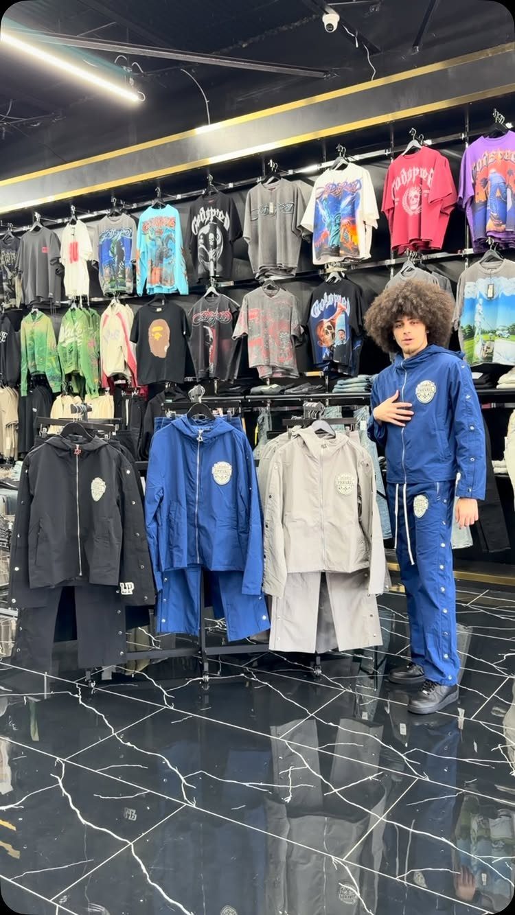 A person with an afro stands among racks of graphic t-shirts and various colored tracksuits.