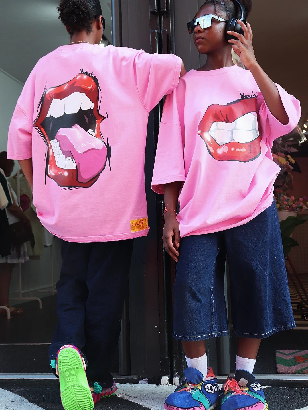 Two people wearing oversized pink graphic t-shirts and dark denim shorts standing outdoors.