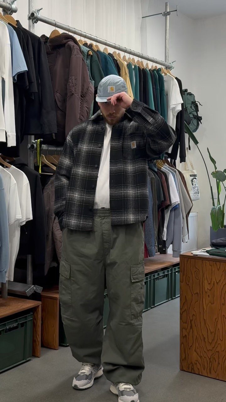 Man wearing a plaid shirt, light blue cap, and olive green cargo pants standing in a clothing store.