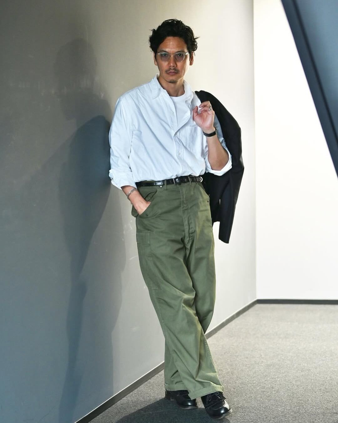 Man in white shirt and olive green cargo pants leaning against a grey wall.