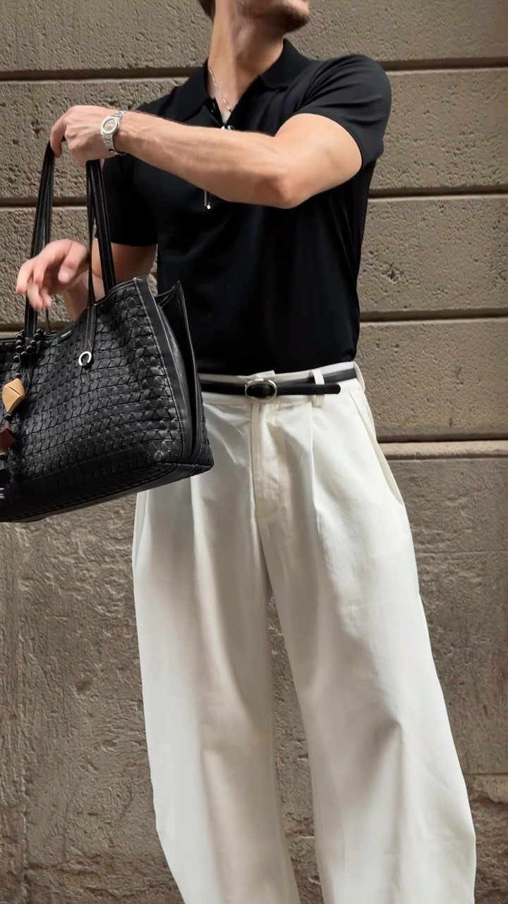 Man in black polo and white trousers holding a large black woven handbag.