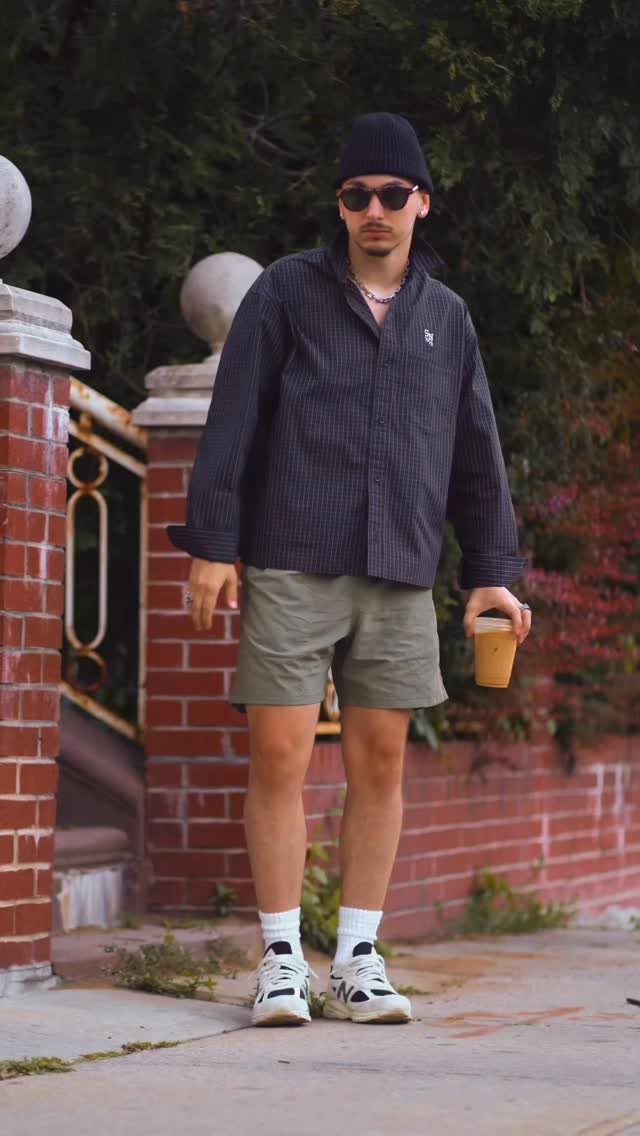 A man walking in a dark striped shirt, olive shorts, and white sneakers.