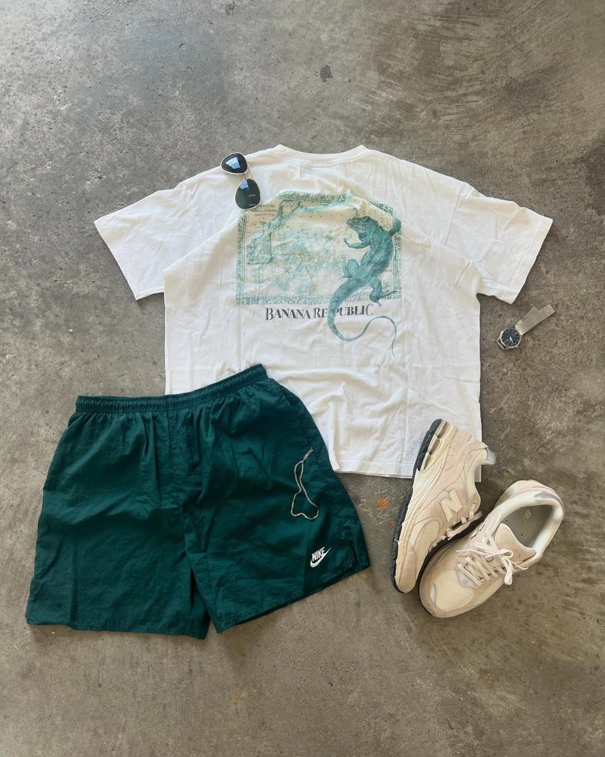 White graphic t-shirt, green shorts, beige sneakers, sunglasses, and a watch laid out on concrete.