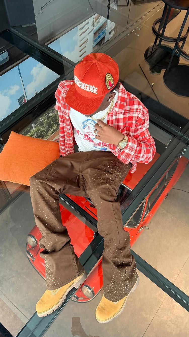 A man wearing a red cap, plaid shirt, and brown trousers sitting on a glass floor.