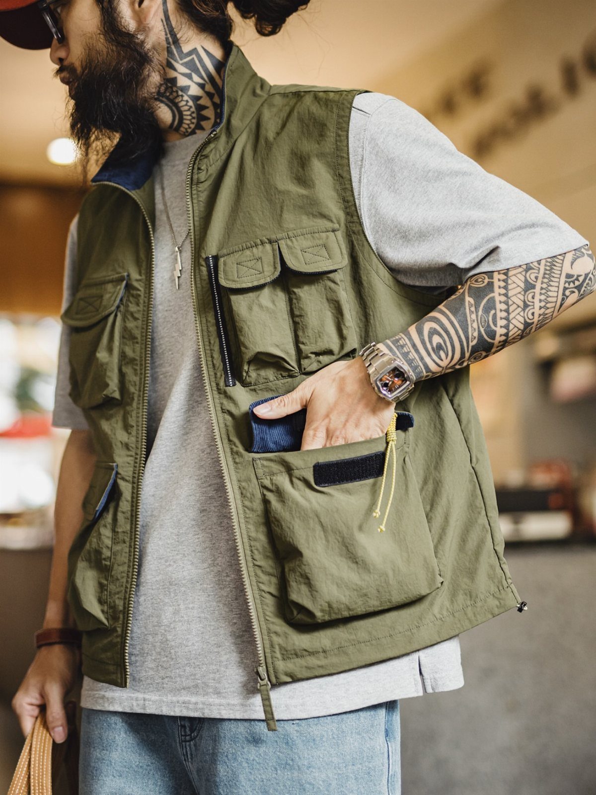 A man wearing an olive green utility vest with multiple pockets over a grey t-shirt.