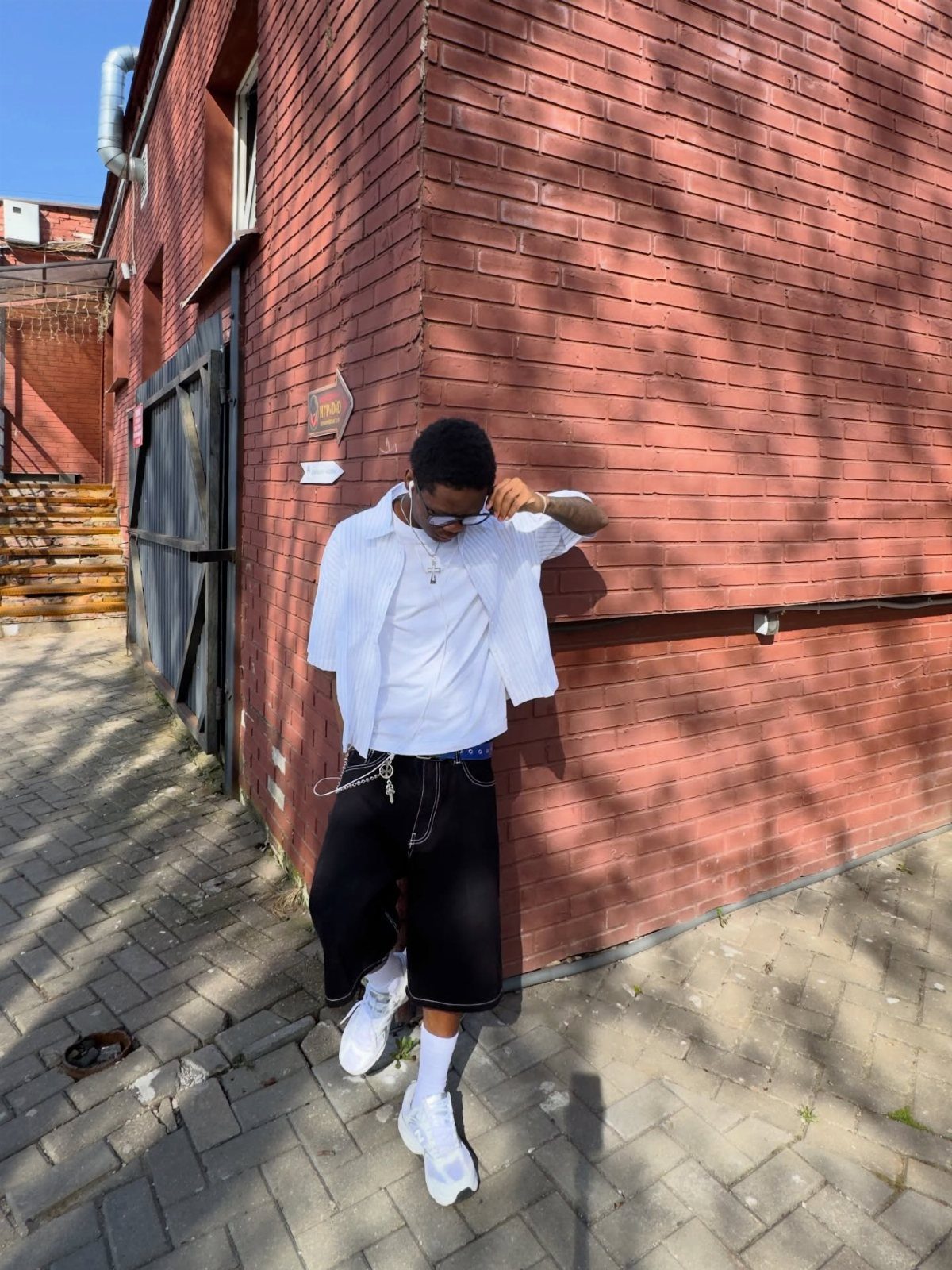 Man in white shirt and black shorts leaning against a red brick wall outdoors.