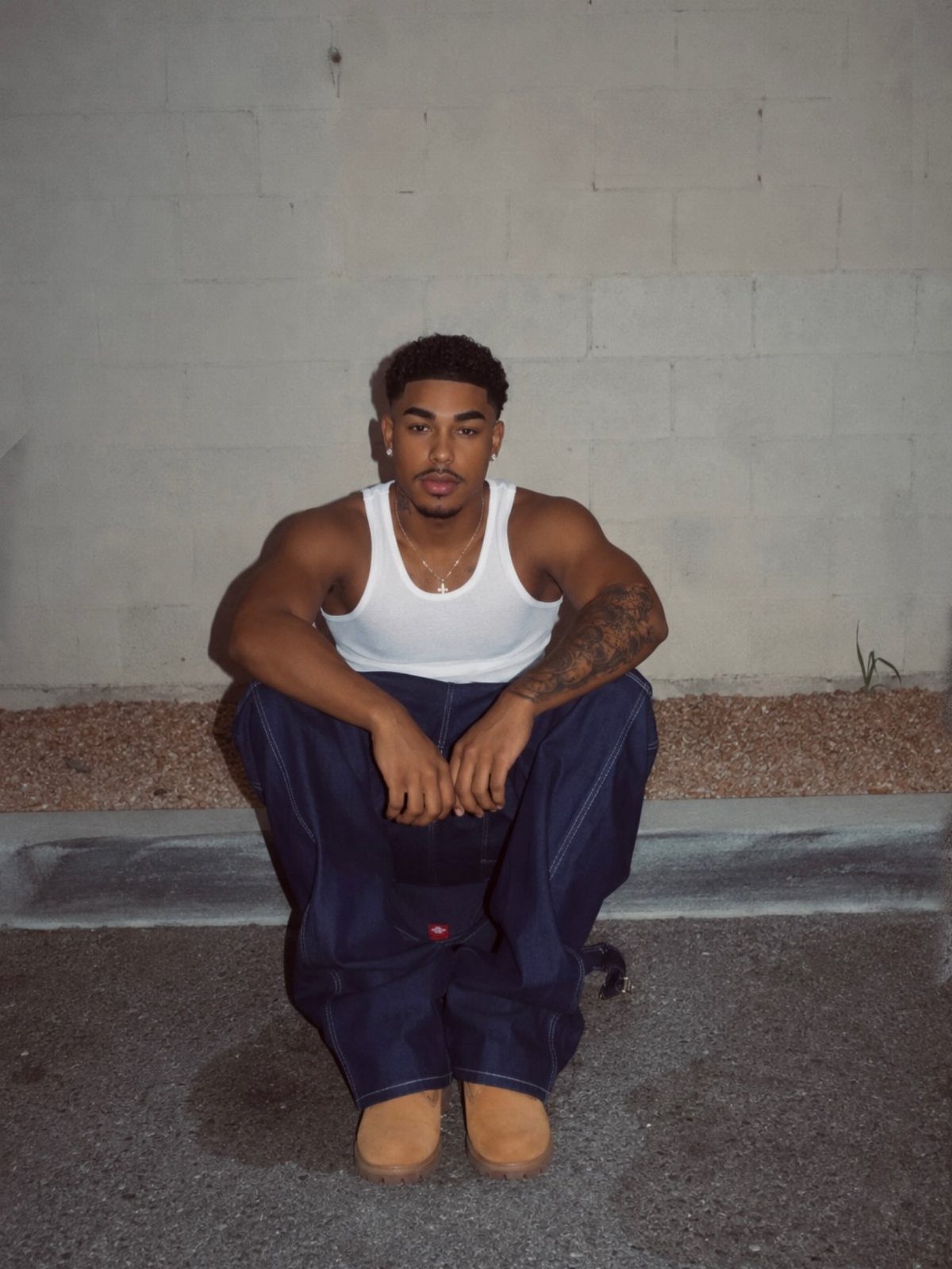 A young man in a white tank top and blue trousers sitting against a concrete wall.