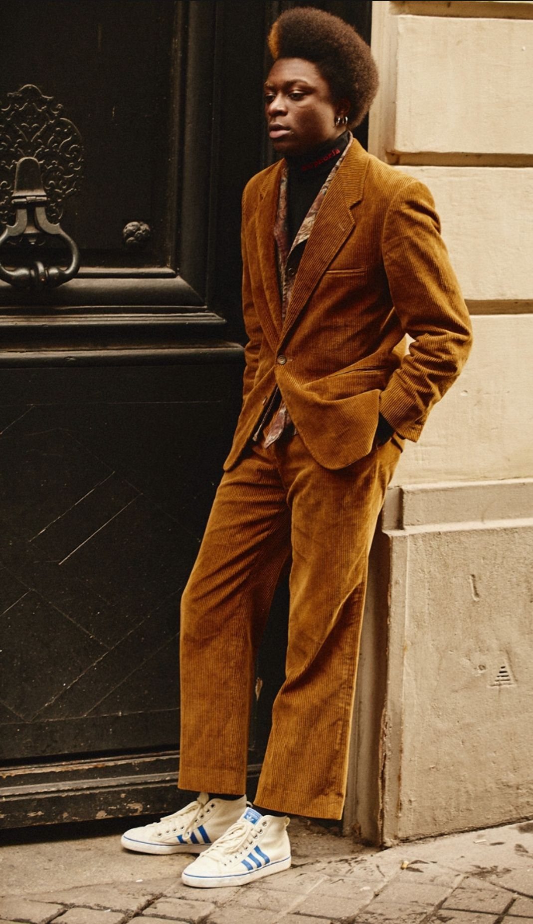A person wearing a brown corduroy suit and white sneakers standing by a dark door.
