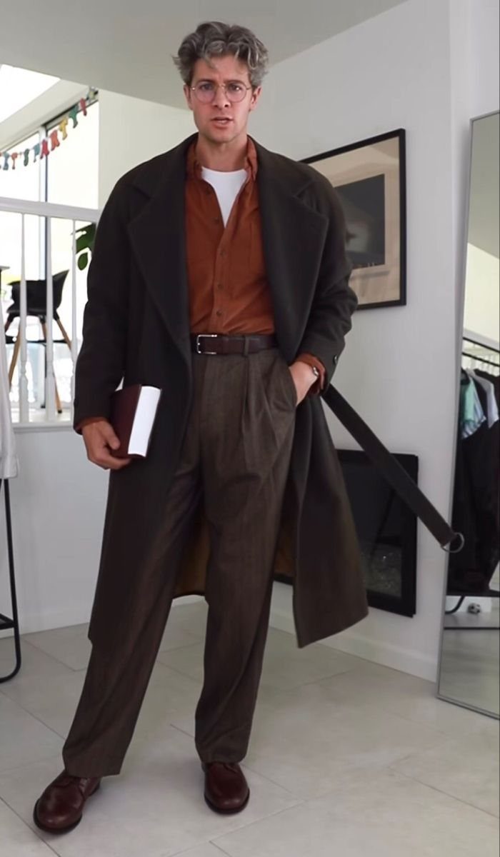 A man wearing a long olive coat, rust shirt, and brown trousers holding a book.