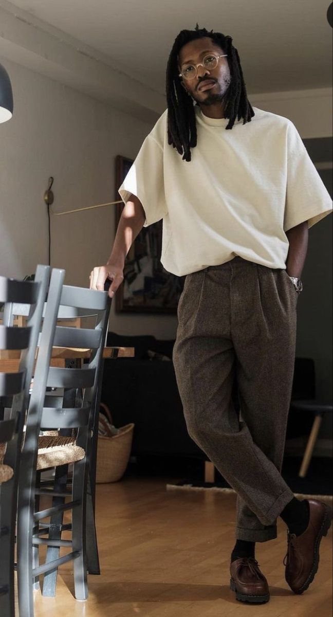 A man wearing a cream t-shirt and brown pleated trousers standing in a living room.