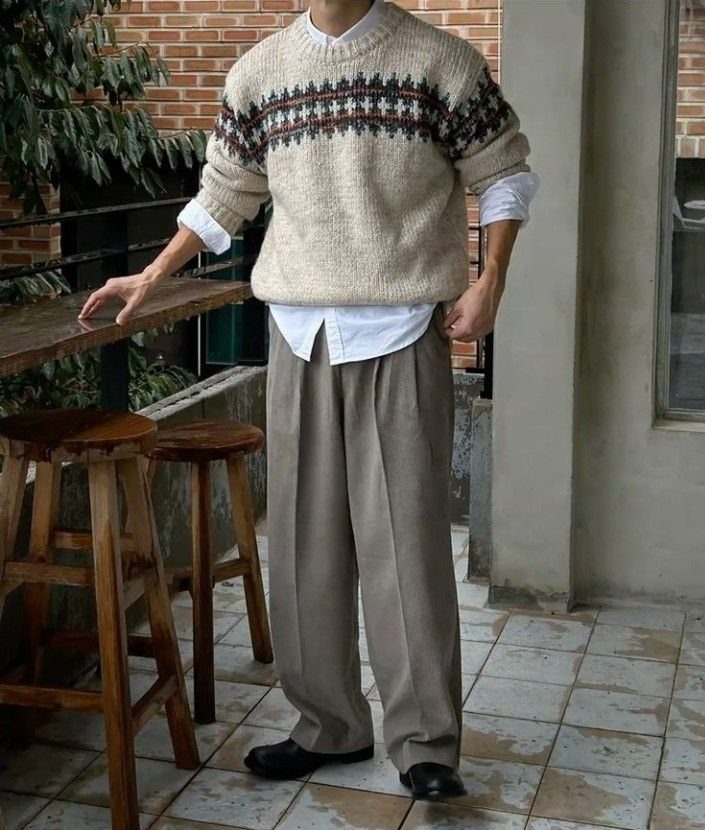 A person wearing a patterned cream sweater, white shirt, and beige trousers standing outdoors.