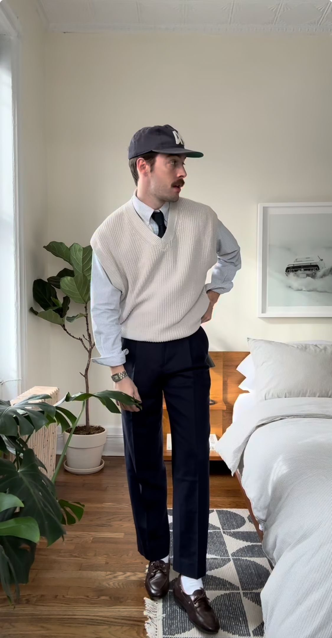 A man posing in a light blue button-down shirt, tie, cream sweater vest, and navy trousers.