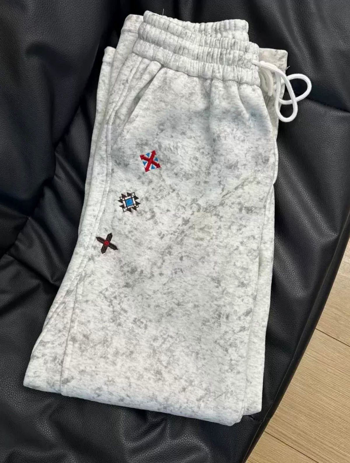 Light grey and white tie-dye sweatpants with small embroidered patches on a black surface.
