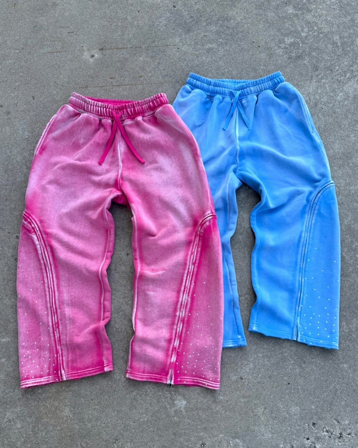 Two pairs of tie-dyed sweatpants, one pink and one blue, lying on concrete.