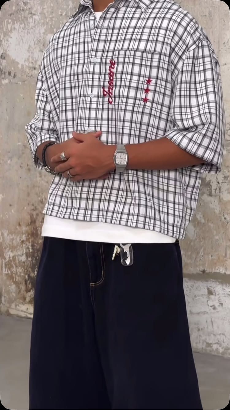 Man wearing an oversized checkered shirt and dark baggy trousers standing against a wall.