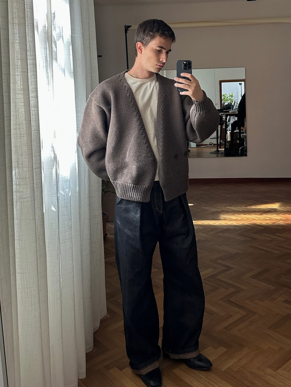 A man posing in a mirror wearing an oversized brown cardigan and wide-leg dark jeans.