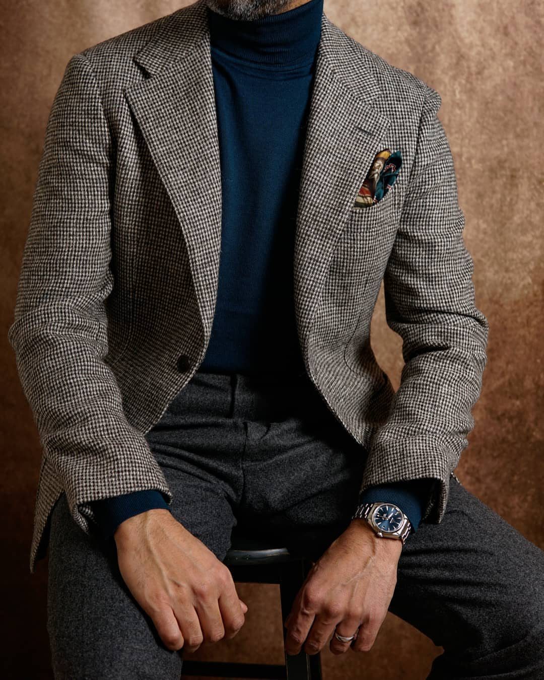 Man wearing a blue turtleneck sweater under a patterned grey blazer and dark trousers.