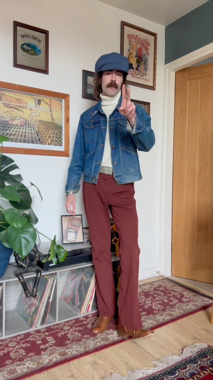 Man wearing a white turtleneck, blue denim jacket, and reddish-brown trousers standing in a room.