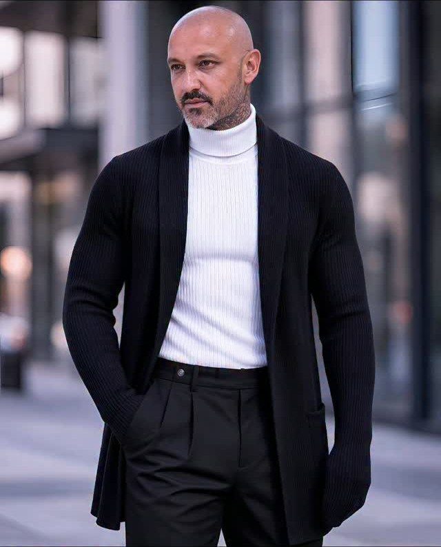 A man wearing a white ribbed turtleneck sweater and a black cardigan outdoors.