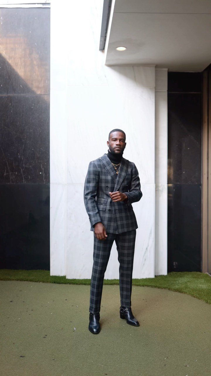 A man in a dark plaid suit and black turtleneck standing against a marble wall.