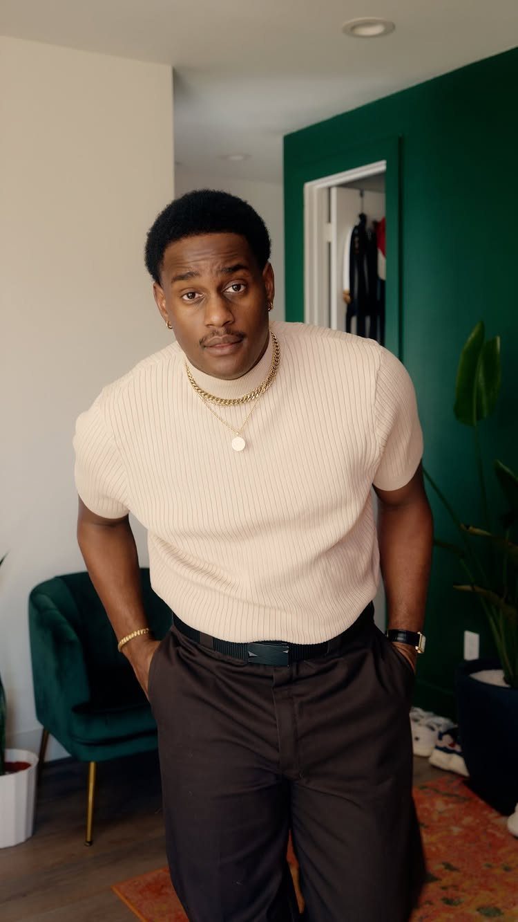A man wearing a cream ribbed short-sleeved mock turtleneck shirt and dark trousers.