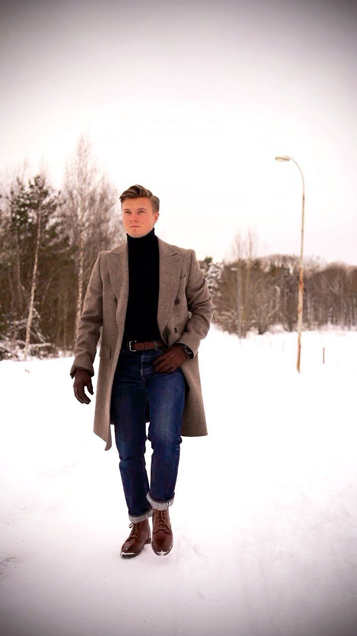 Man in a black turtleneck, grey coat, and blue jeans walking through snow.