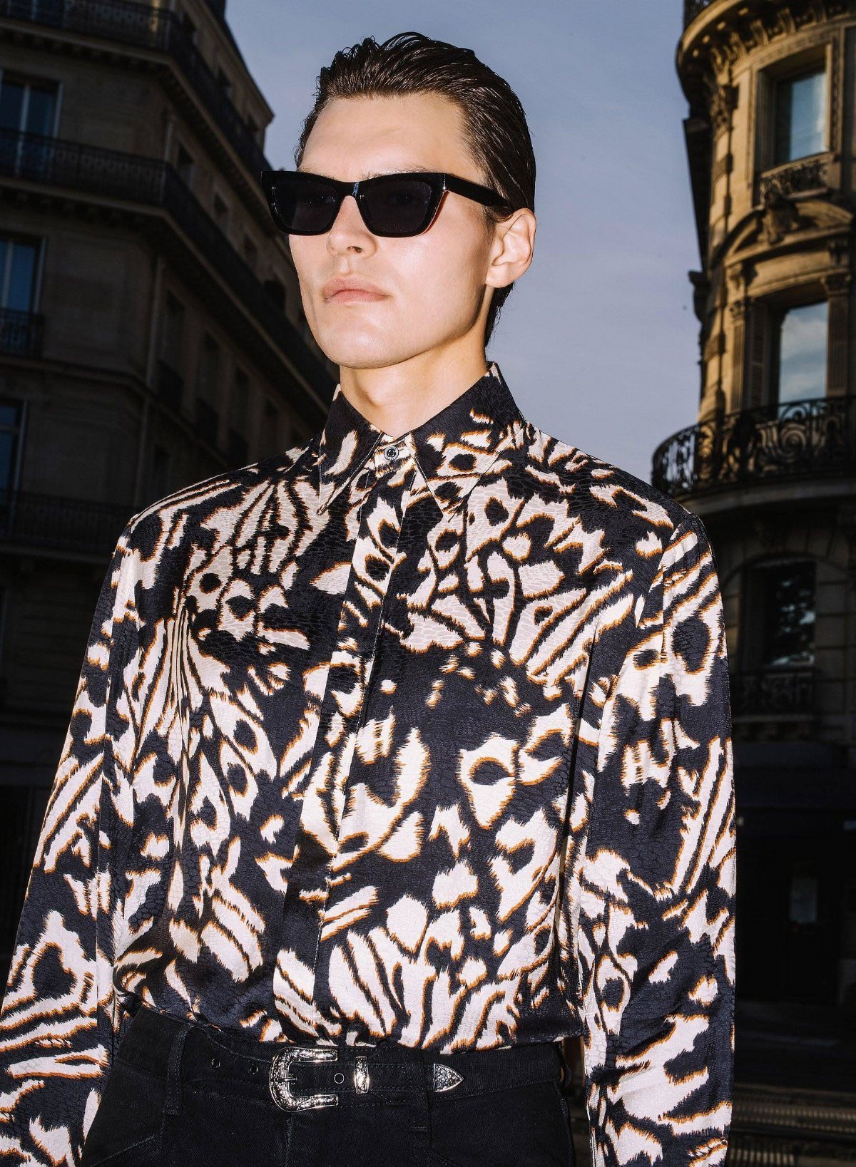 Man in sunglasses wearing a patterned black and cream long-sleeved shirt outdoors.