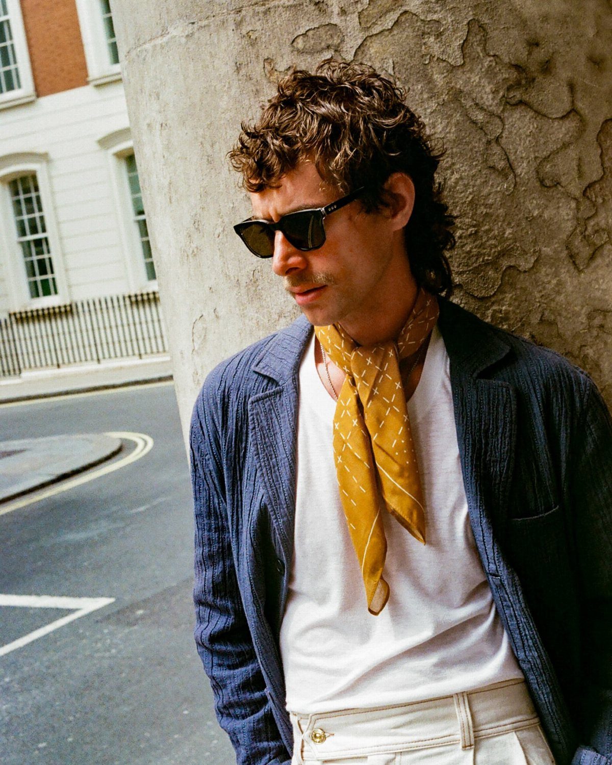 A man wearing a yellow patterned bandana tied around his neck while leaning against a pillar.