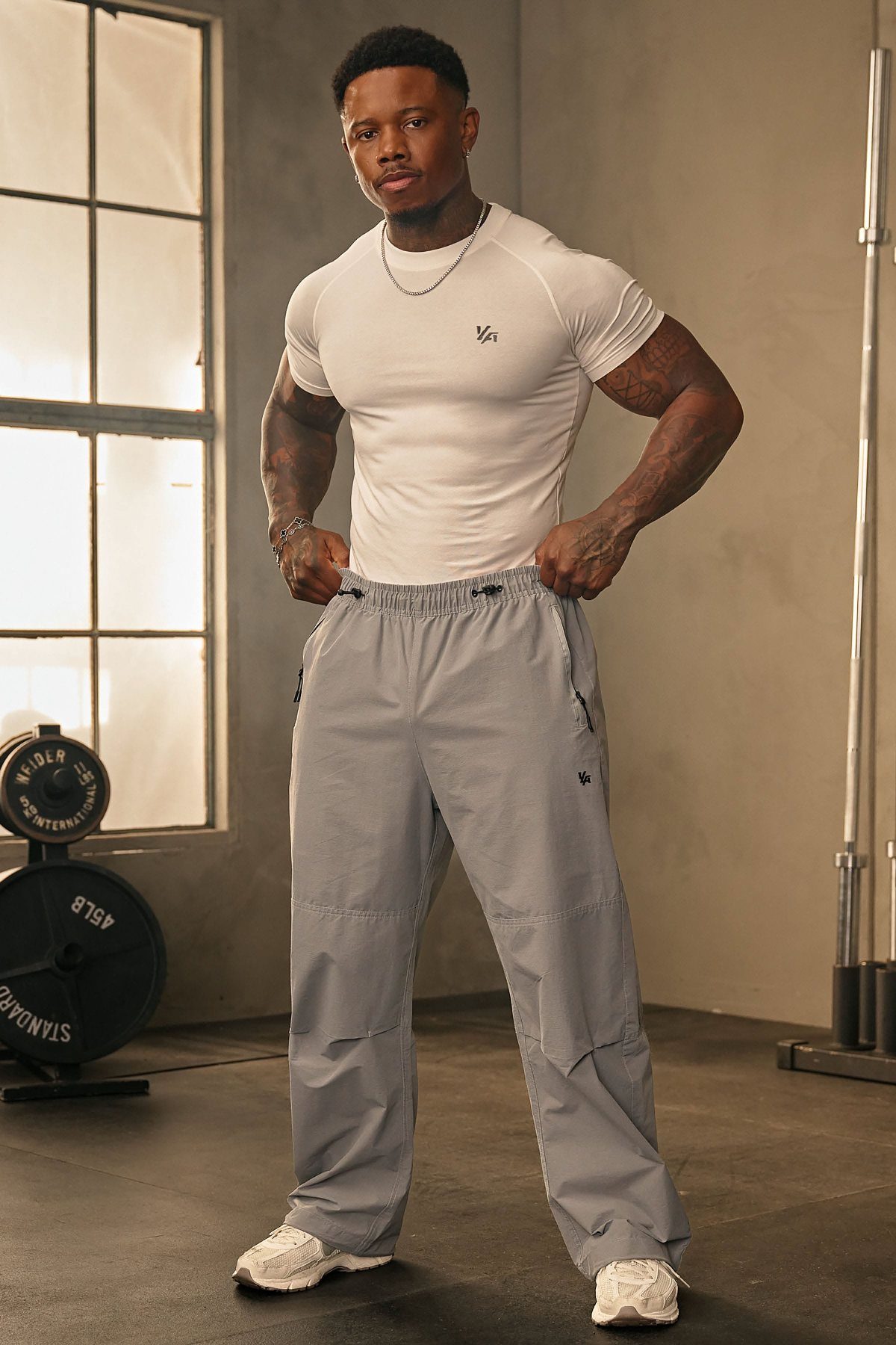 A muscular man wearing a white athletic shirt and grey joggers posing in a gym.