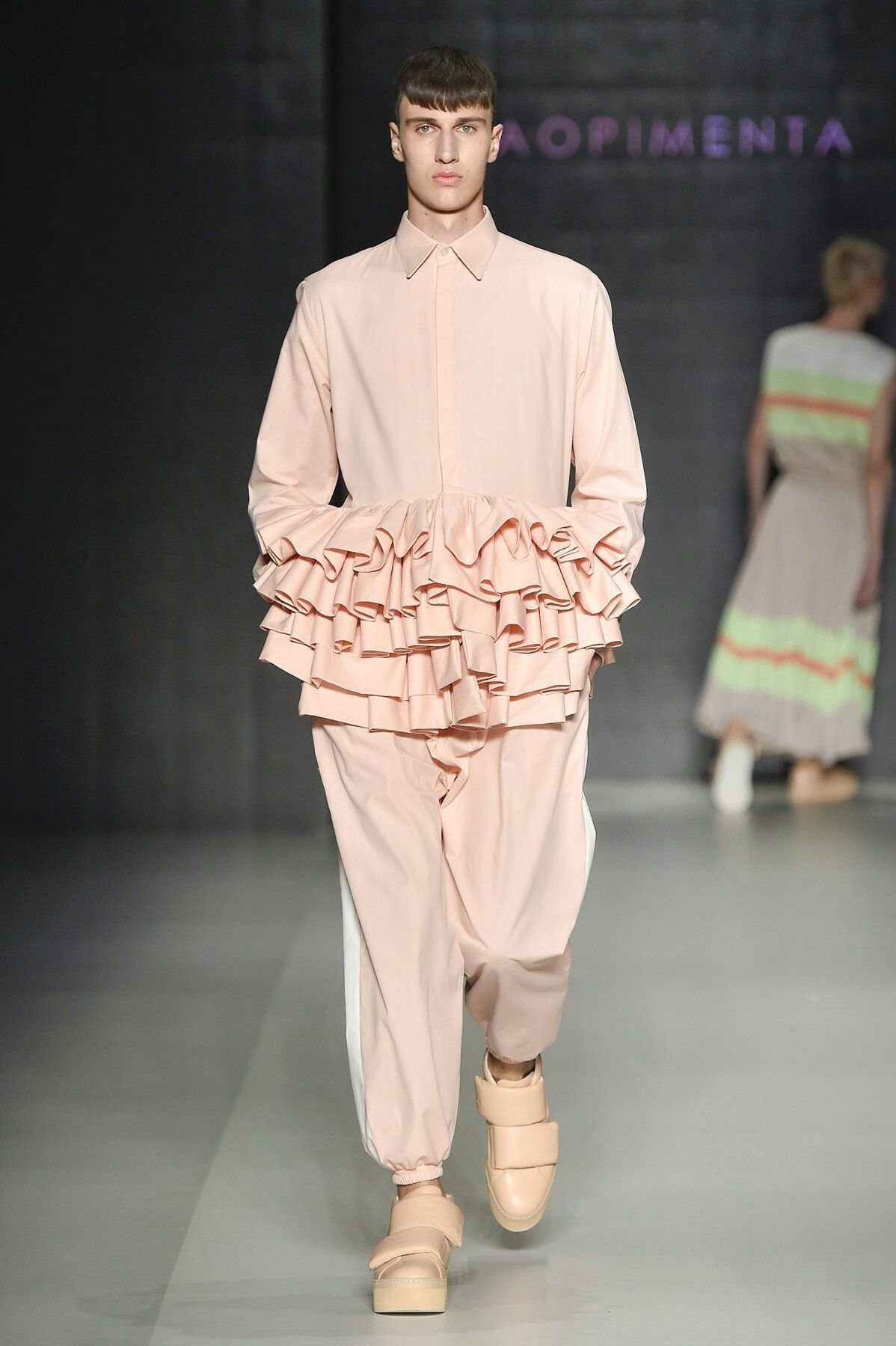 Male model walking a runway in a pink ruffled shirt and matching trousers.