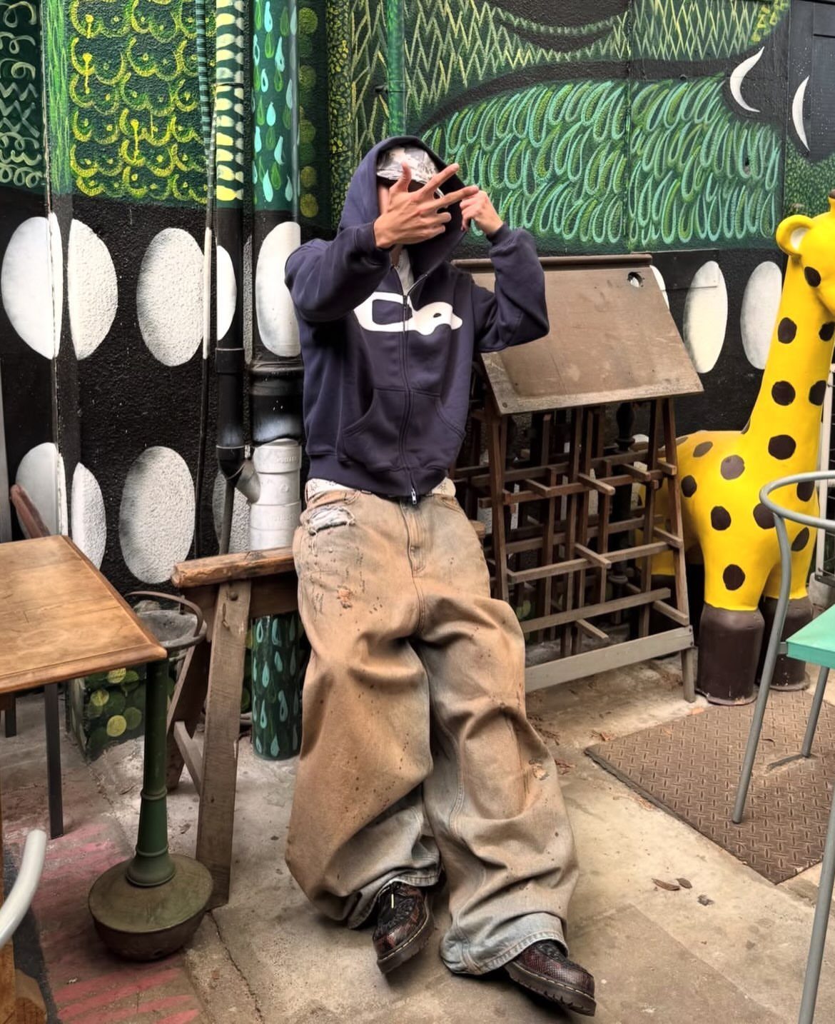 Person wearing an oversized dark hoodie and baggy tan trousers against a graffiti wall.
