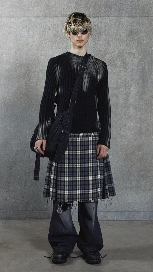 A person wearing a black ribbed top, plaid kilt, and wide-leg trousers against a grey wall.