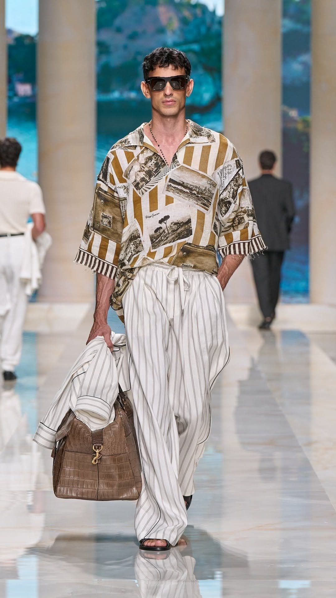 A male model walking a runway in a patterned shirt and striped wide-leg trousers.