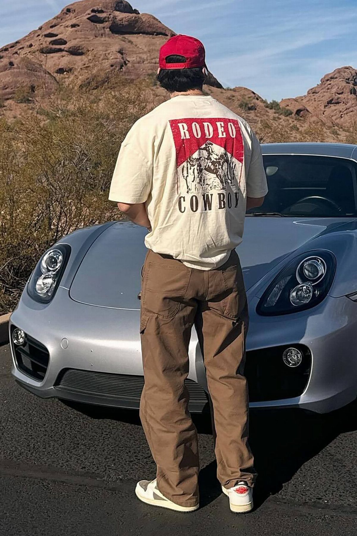 A person wearing a cream graphic rodeo cowboy t-shirt and brown cargo pants standing by a car.
