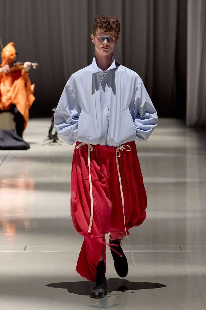 A male model walking a runway in oversized blue shirt and voluminous red trousers.