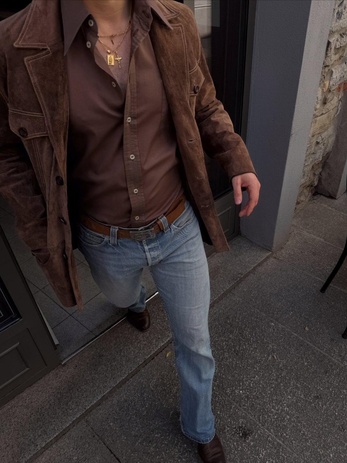 Man wearing a brown suede jacket, brown shirt, and light blue denim jeans standing outdoors.