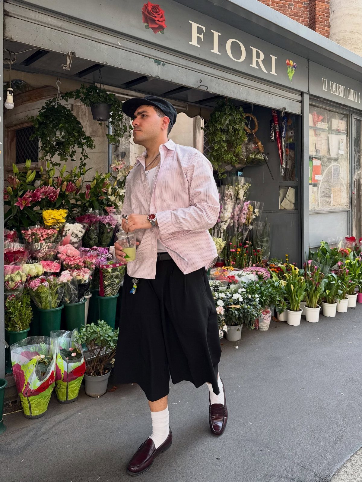 A man in a pink striped shirt, black wide-leg trousers, and loafers standing by a flower shop.