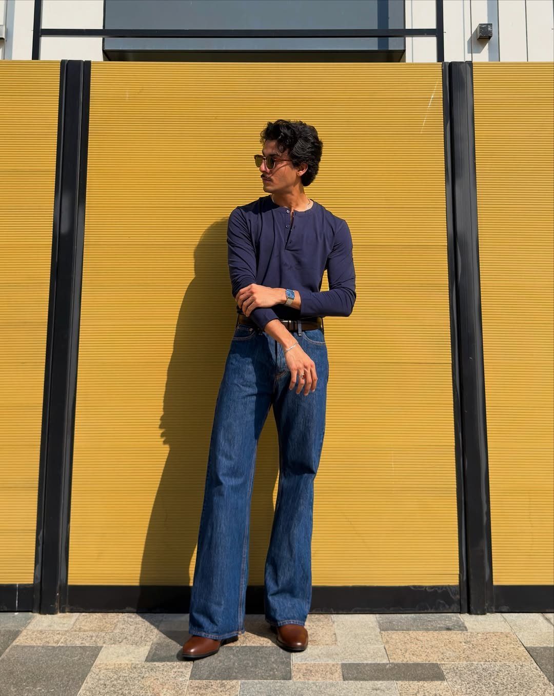 A man posing in blue flared denim jeans and a dark blue long-sleeved shirt against a yellow wall.
